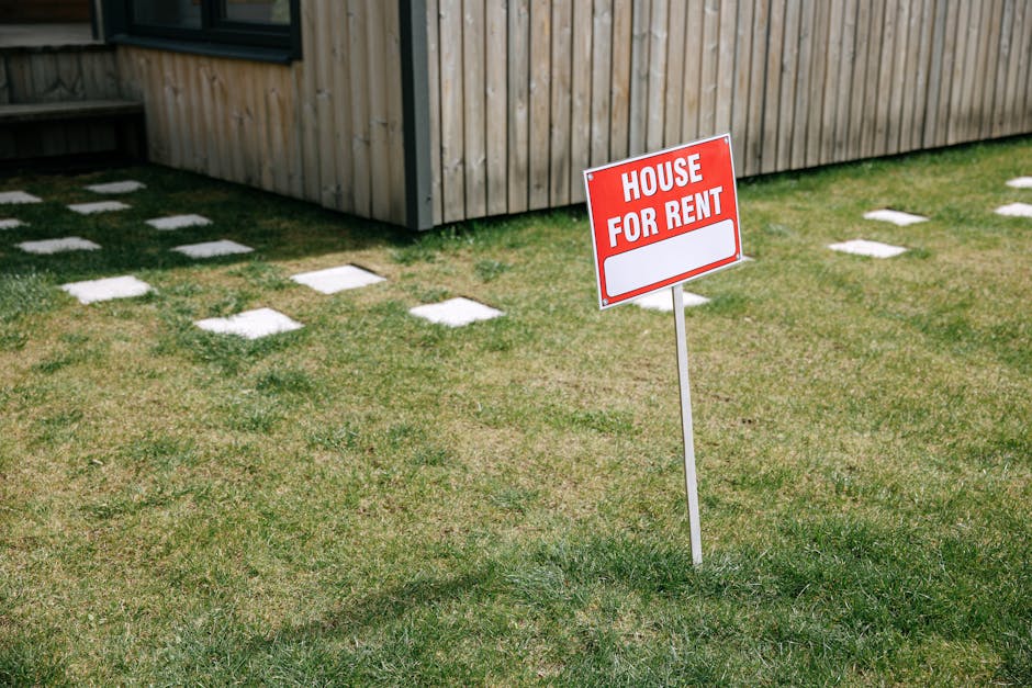 A red  House for Rent  sign stands on a grassy lawn beside a wooden house exterior.